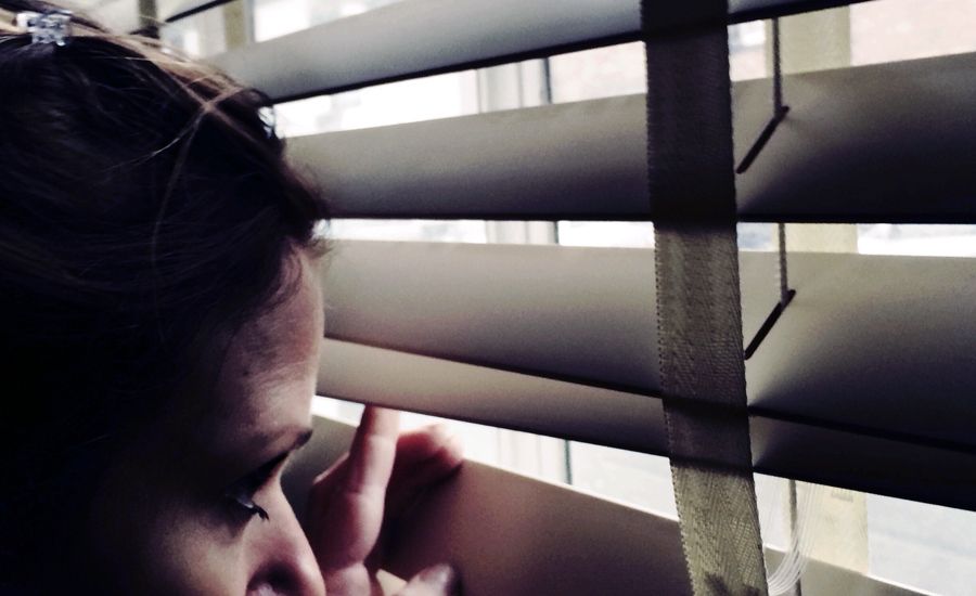 A woman peers anxiously out through her closed blinds.